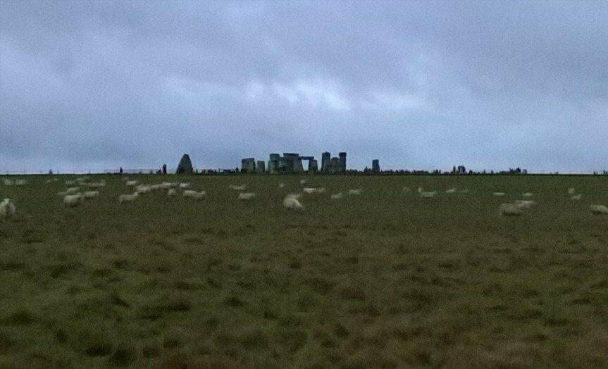 Auto-generated description: A flock of sheep grazes in a grassy field beneath a cloudy sky, with Stonehenge visible in the background.