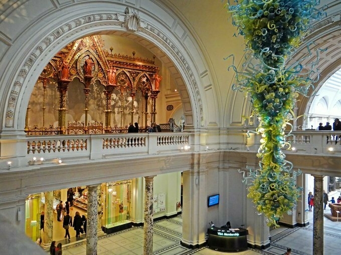Auto-generated description: A grand interior features ornate architectural details, an elegant chandelier, and an information desk with visitors exploring the space.