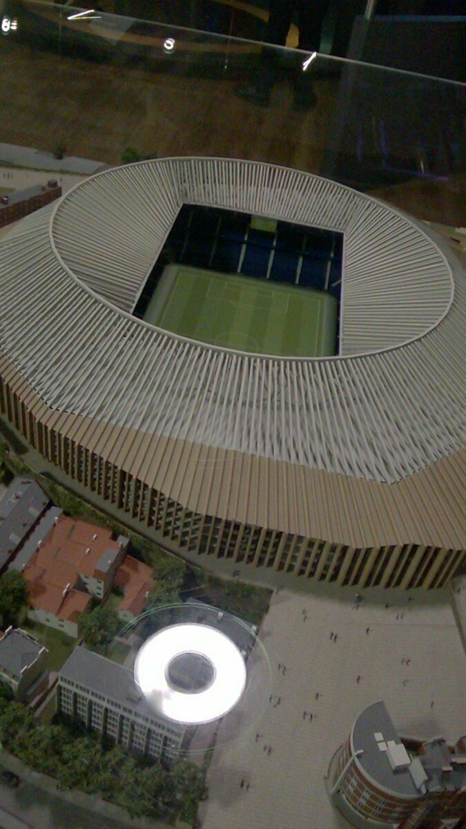 Auto-generated description: A scale model of a modern football stadium with a distinctive circular roof design is displayed, surrounded by miniature buildings and trees.