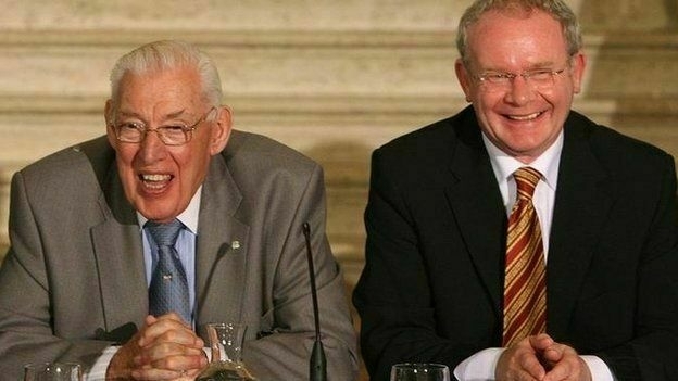 Auto-generated description: Two men are sitting side by side, both smiling, in formal attire with a wooden backdrop behind them. The two men are Martin McGuiness and Ian Paisley and I still can't believe it.
