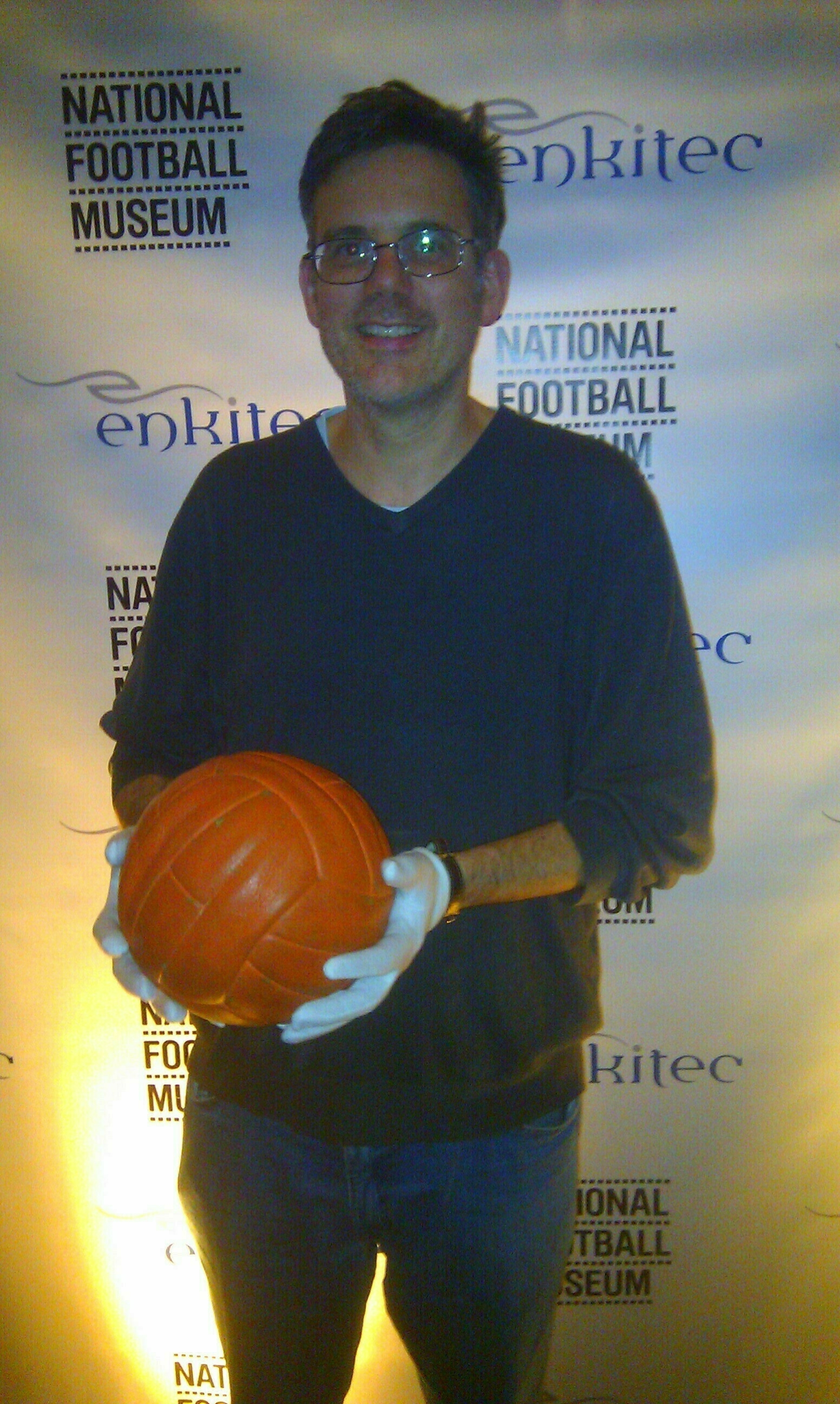 Auto-generated description: A person is holding an orange basketball in front of a backdrop featuring the logos National Football Museum and enkitec.