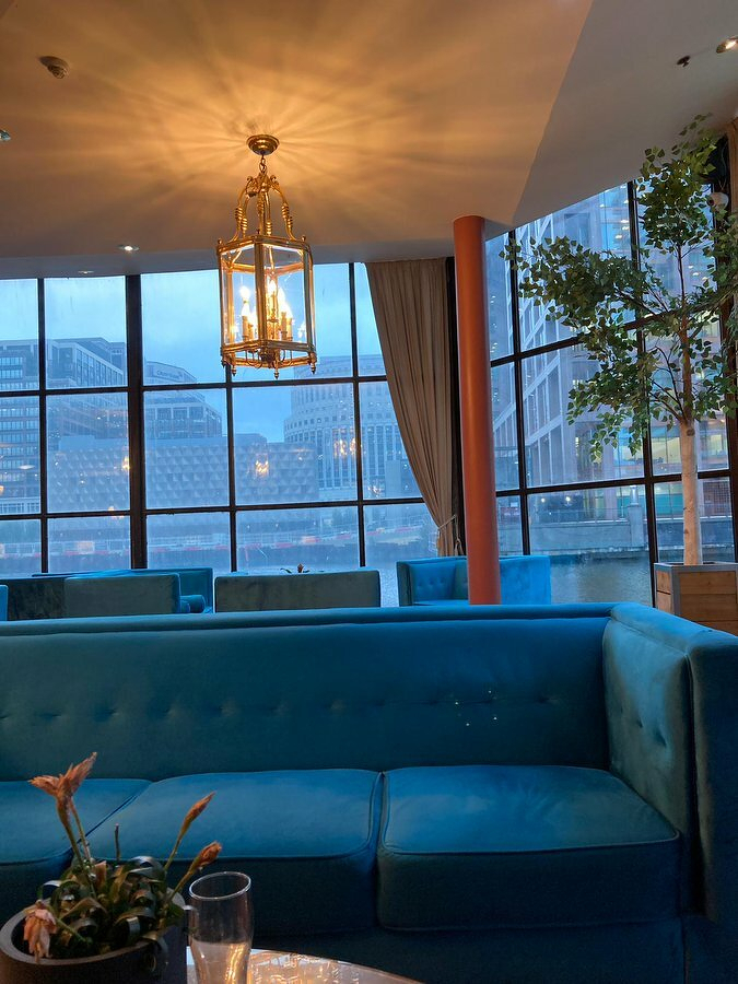 Auto-generated description: A cozy indoor lounge with a blue sofa is set against large windows offering a view of modern buildings across the water.