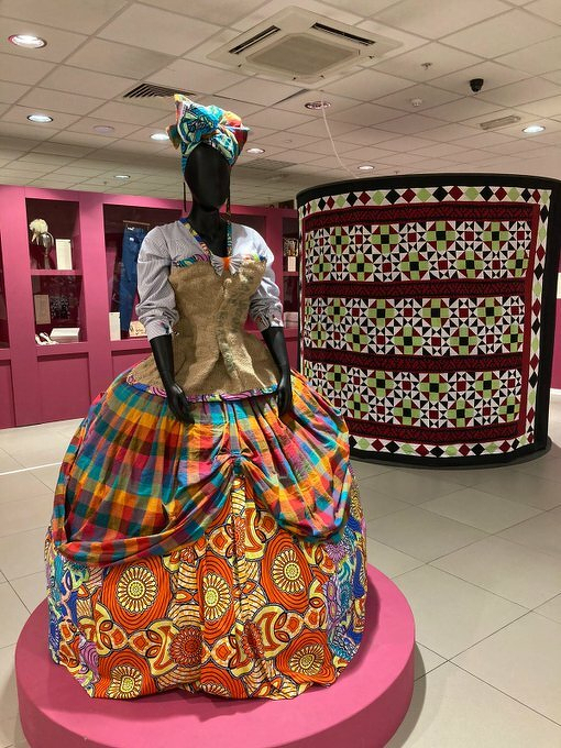 Auto-generated description: A mannequin is dressed in a colorful, traditional outfit with a matching headwrap, set on a pink platform in a room with patterned textiles.