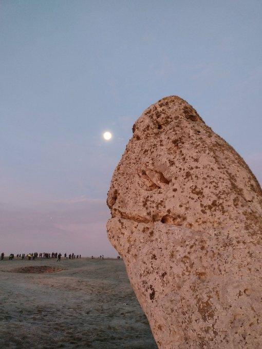 Auto-generated description: A rock formation resembling a face is lit by the moon, with a group of people visible in the distance on a barren landscape.