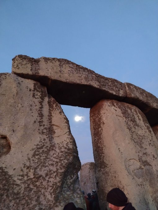 Auto-generated description: A partial view of Stonehenge shows its massive standing stones with the moon visible through a gap.