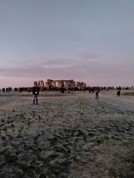 Auto-generated description: Stonehenge is surrounded by a large group of people on a frosty morning under a pink sky.