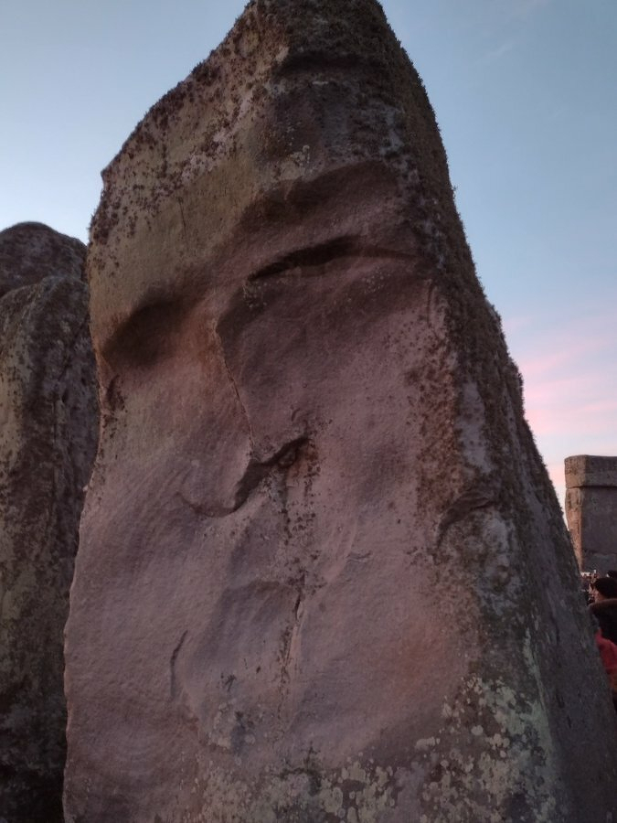 Auto-generated description: A large, weathered stone with mossy patches exhibits a face-like appearance, set against a twilight sky.