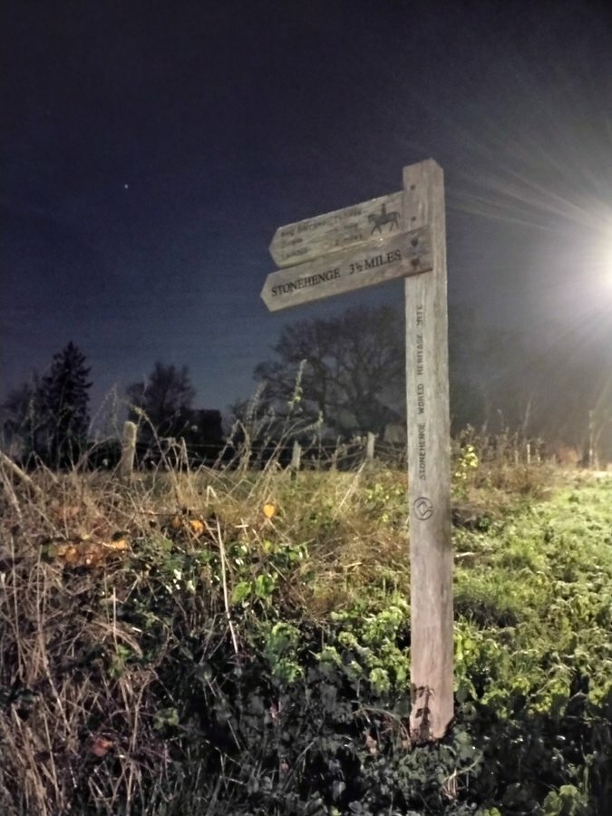 Auto-generated description: A wooden signpost in a grassy area points towards Stonehenge, 3½ miles away, under a night sky with a bright light illuminating the scene.