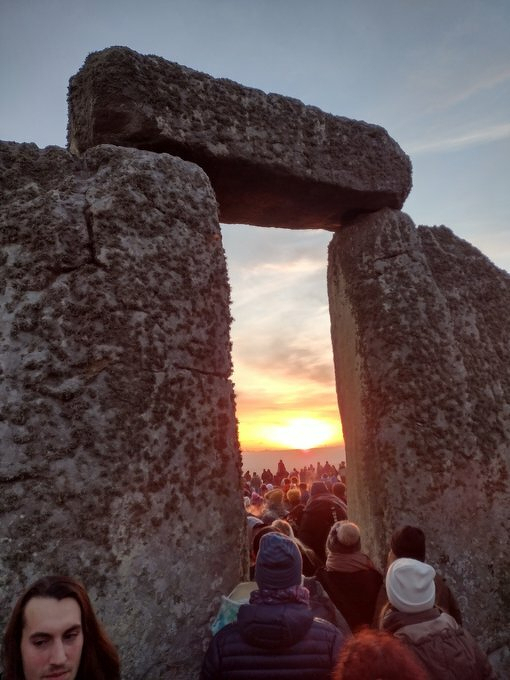 Auto-generated description: Crowds gather at Stonehenge, viewing the sunset framed by its iconic stone structures.
