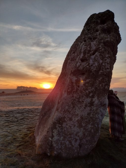 Auto-generated description: A large standing stone is silhouetted against a colorful sunset sky in a grassy landscape.