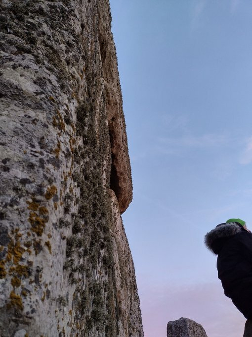 Auto-generated description: A large textured rock formation stands against a blue sky with a hint of a person wearing a winter coat visible at the edge.
